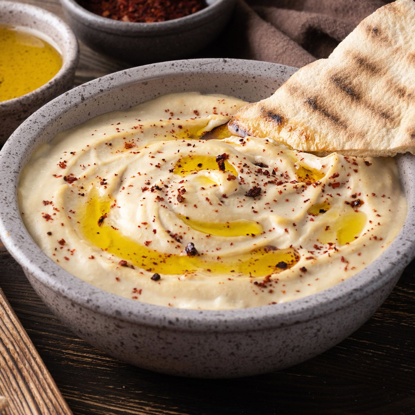 A close-up of a speckled gray ceramic bowl filled with creamy hummus, topped with a swirl of golden olive oil and red chili flakes, with a piece of grilled pita bread dipping into the sauce. Small bowls of seasoning and oil are blurred in the background on a dark wooden table.