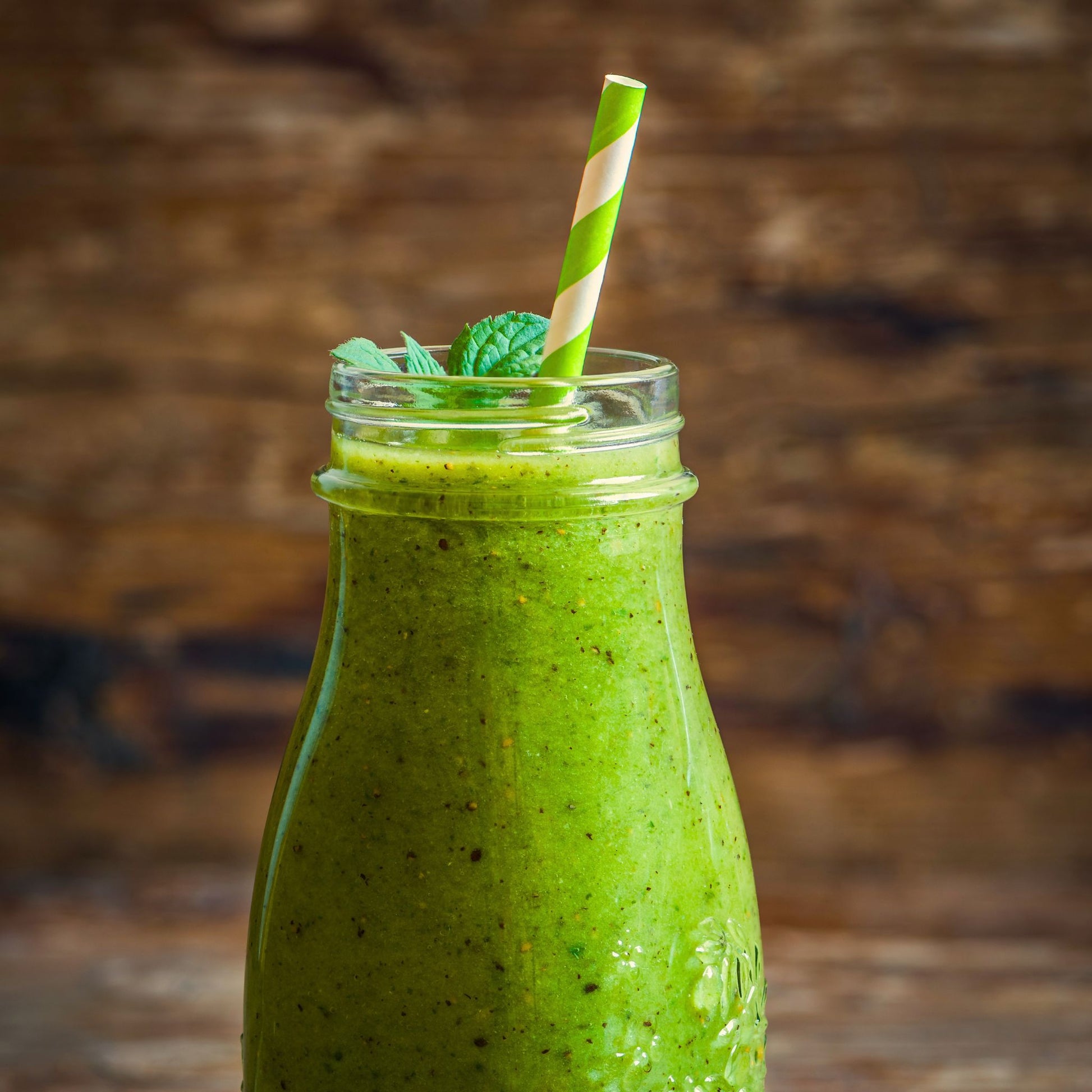 A green smoothie in a clear glass bottle with a striped green and white paper straw, garnished with mint leaves, set against a rustic dark wood background.